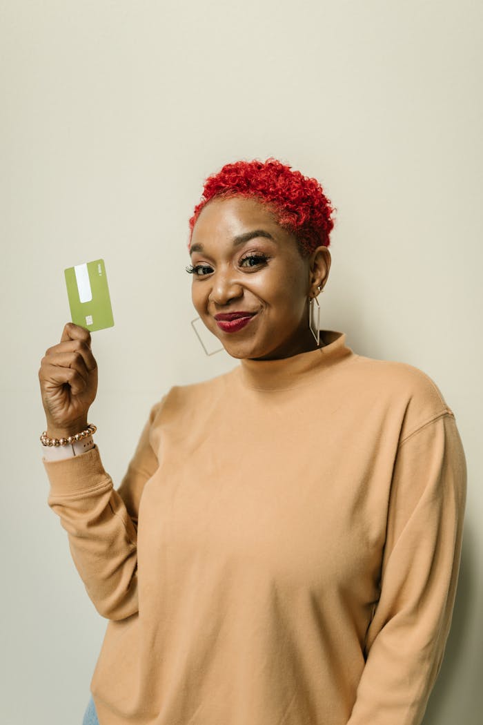 African woman in brown sweater, smiling and holding a green credit card. Vibrant and friendly portrait indoors.